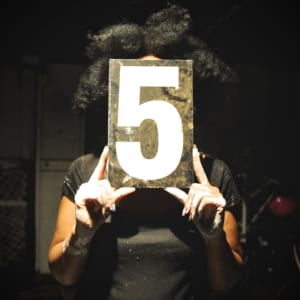 Femme Space key image person holding the number 5 covering face