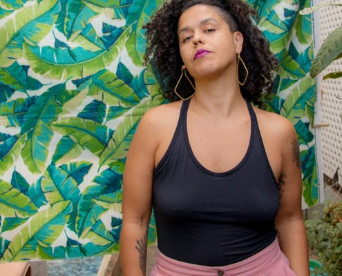 Curly haired brown skin person with pink pants, black leotard, hoop earrings standing against a colorful background