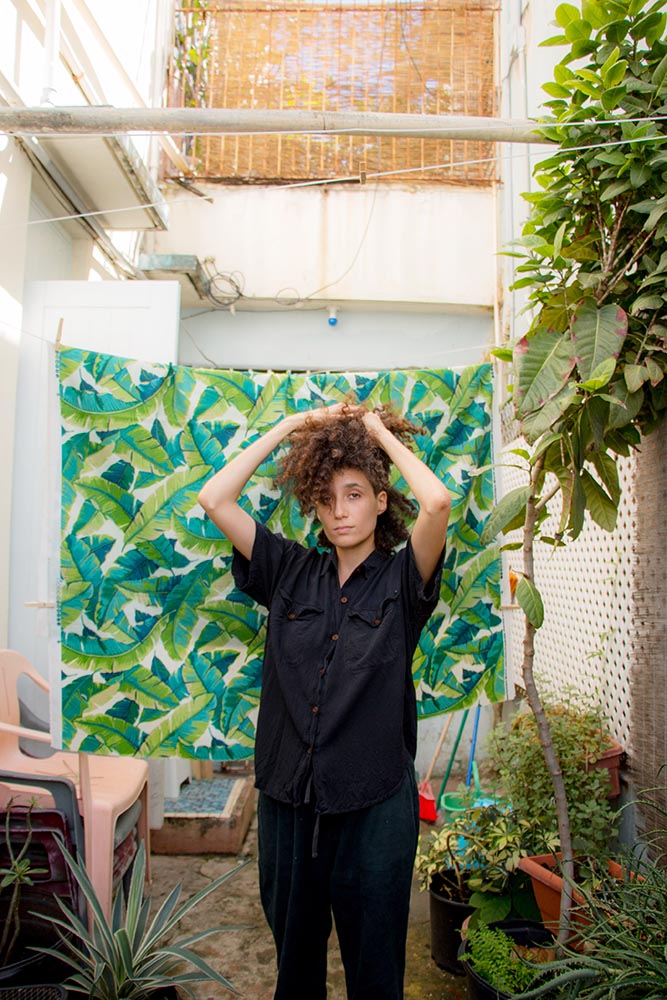 Brown curly haired person holding their hair up in black short sleeved jumpsuit standing against backdrop of green leaves in an outdoor space