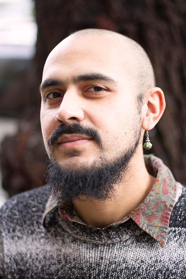 Bald headed person with beard, mustache, and one earring wearing a collared paisley shirt and knitted sweater