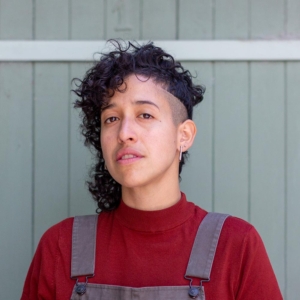 Curly haired person with one side of head shaved wearing a red shirt and overalls