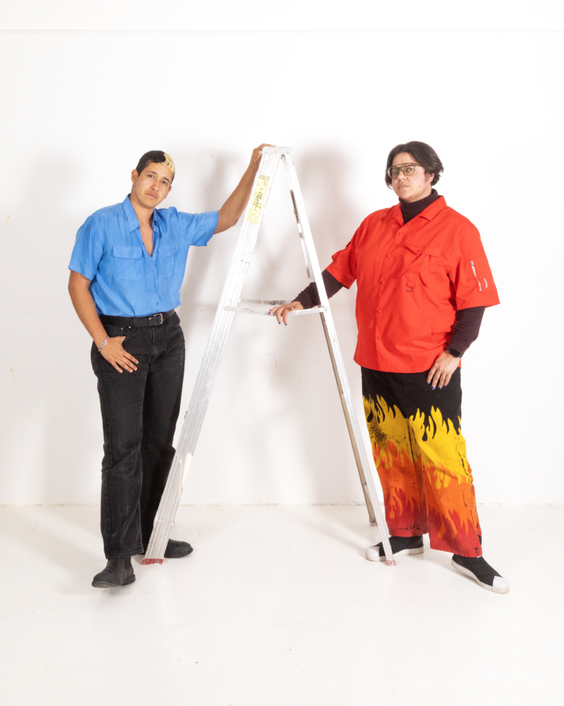 Art Handlxrs project directors Marcel Pardo Ariza (left) and Ambrose Trataris (right) stand on either side of an A-frame ladder against a plain white wall and floor